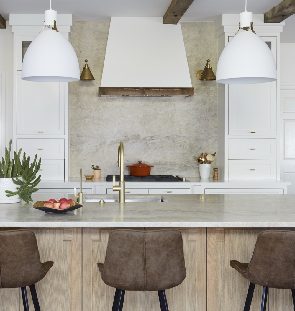 Kitchen island in cinnamon-veined white quartzite on a white oak base, facing a slab quartzite range wall with brass sconces and a plaster hood, by Martire Custom Homes