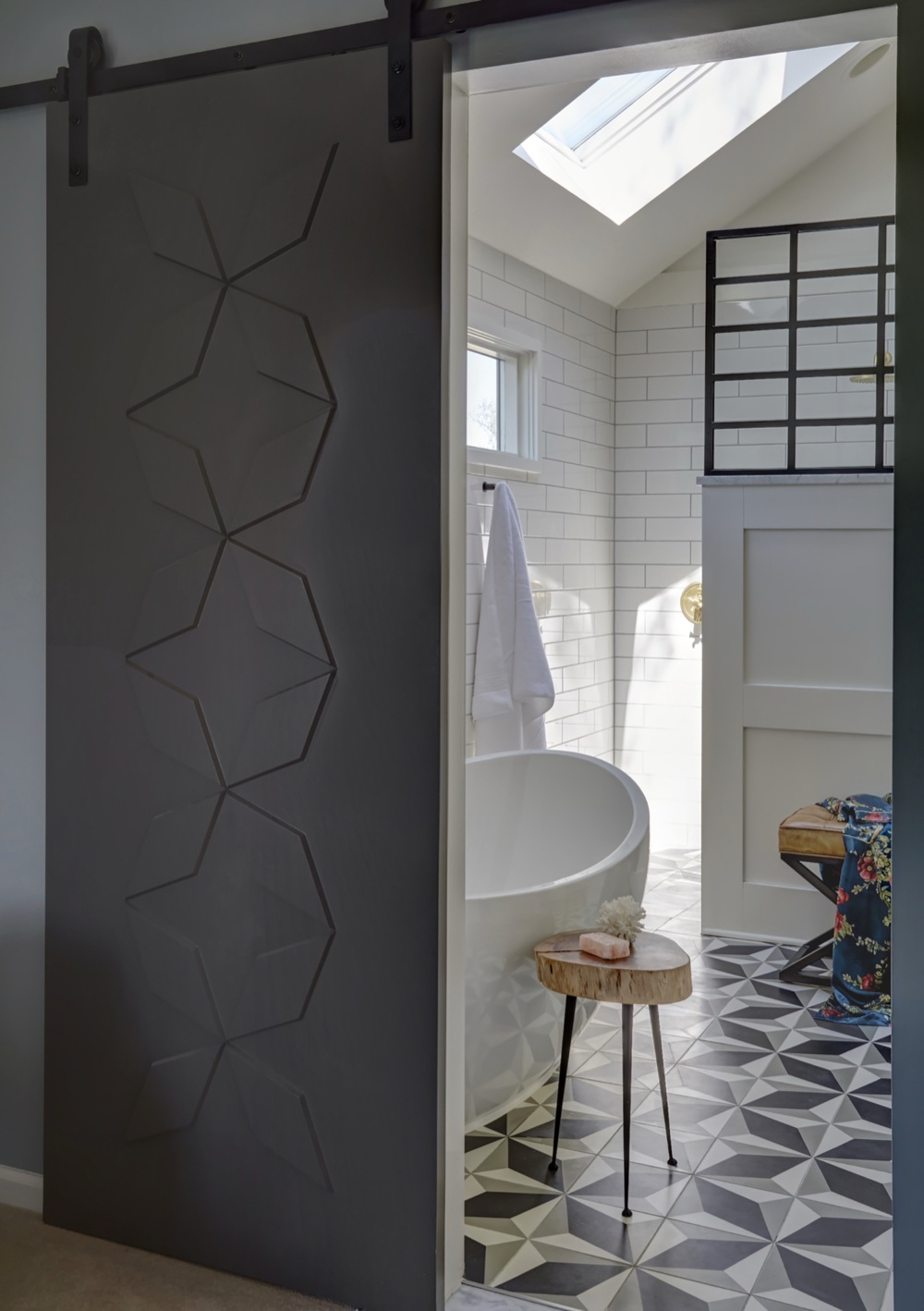 View through a geometric-paneled sliding door into the primary ensuite with freestanding tub, white subway tile, steel-framed shower screen and patterned cement floor, by Martire Custom Homes