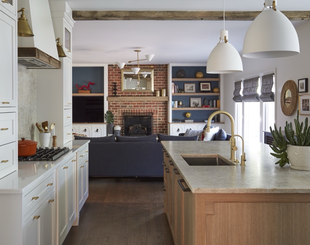 Open-plan kitchen with white oak island and cinnamon quartzite counters flowing into a family room with exposed brick fireplace, navy sectional and built-in shelving, by Martire Custom Homes