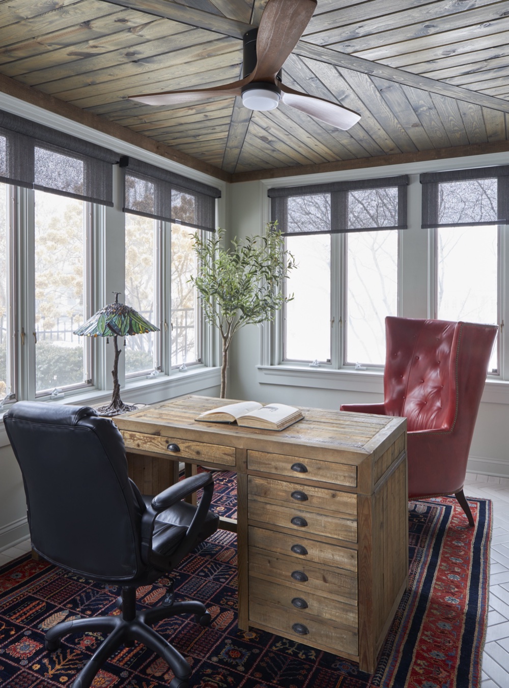 Sunlit gentleman's office with rustic reclaimed-wood desk, red tufted leather wing chair, stained-glass lamp, and wood plank ceiling, by Martire Custom Homes