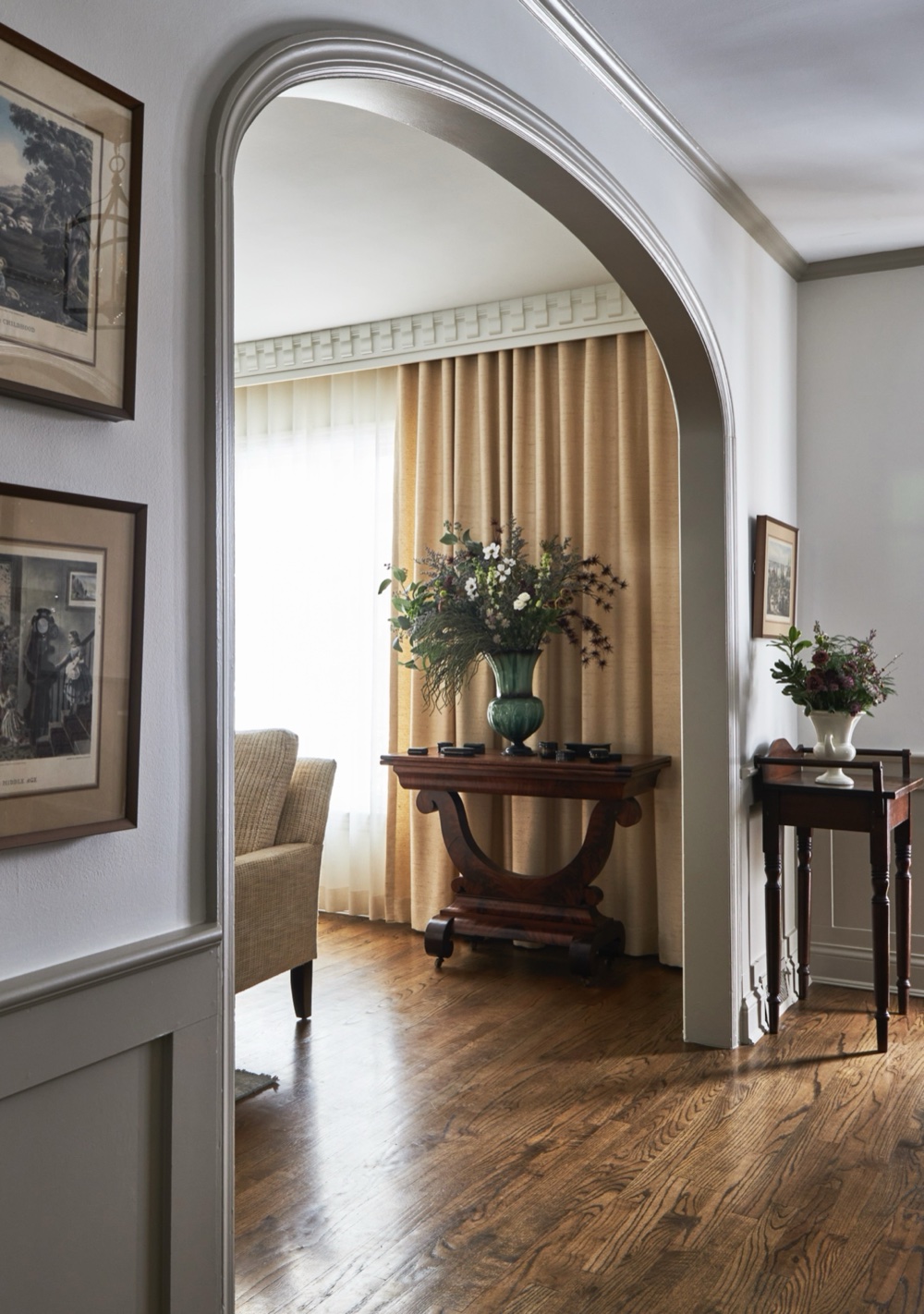 View through an arched foyer opening to an antique console with green glass vase against linen curtains, by Martire Custom Homes