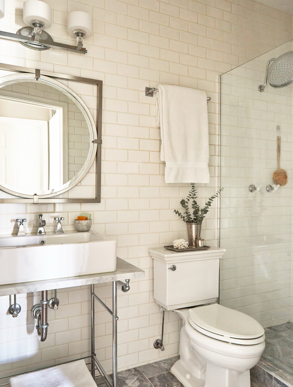 Renovated bath with cream subway tile walls, a marble-topped console sink on chrome legs, round pivot mirror and a glass-enclosed rain shower, by Martire Custom Homes