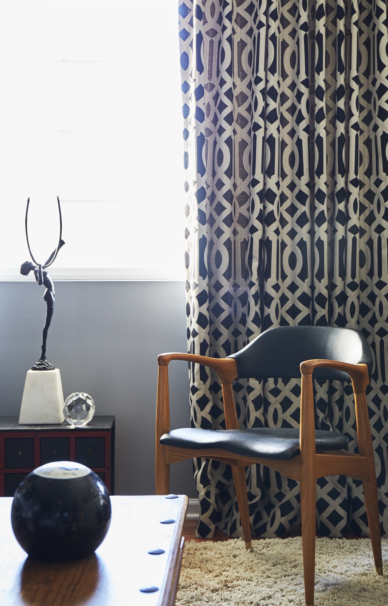 Family room detail with a mid-century oak and black leather armchair, bold trellis-patterned drapery, a bronze figure on marble and a shag rug, by Martire Custom Homes