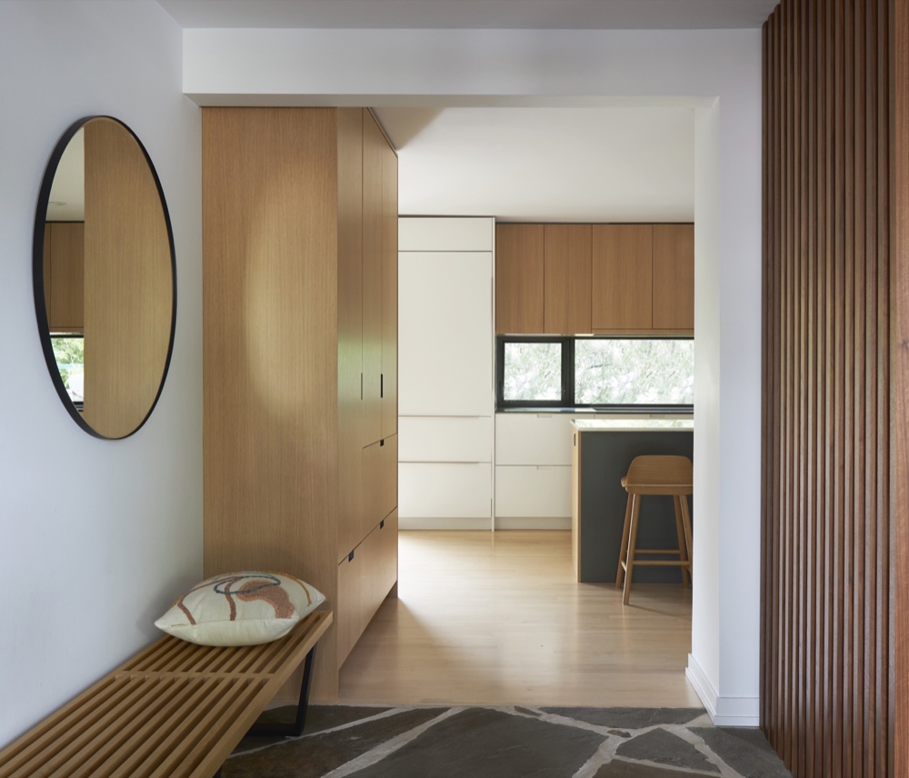 Foyer with slatted wood bench, round mirror and white-oak millwork opening onto a Scandinavian kitchen