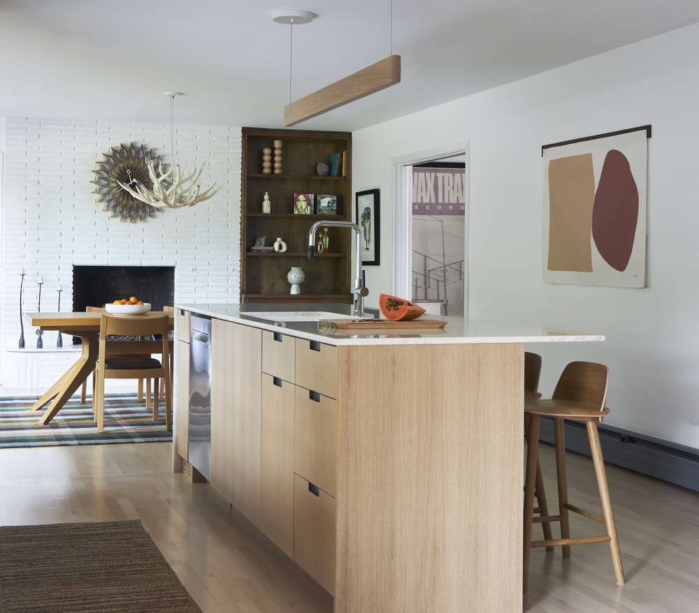 Wide view of white-oak kitchen island with dining table and white brick fireplace beyond
