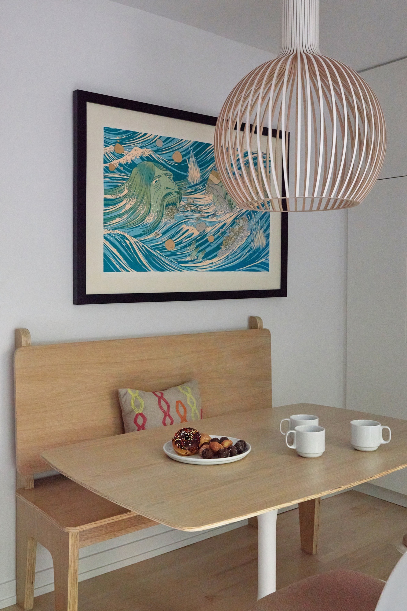 Built-in oak breakfast bench and table beneath a framed wave print and woven pendant