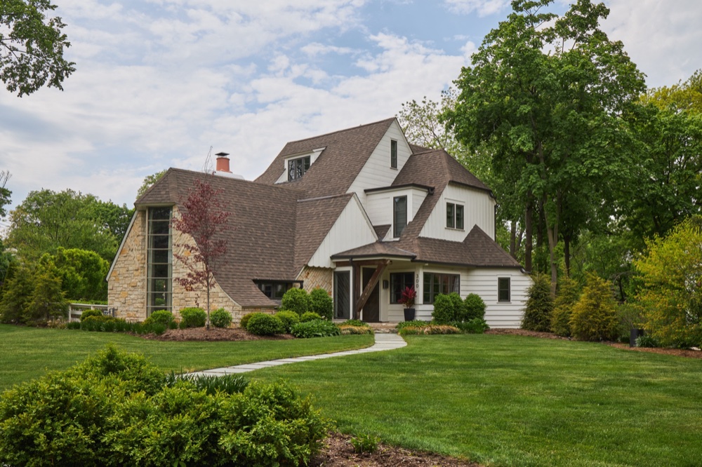 Front exterior of a restored 1920s storybook residence with a sweeping catslide roofline, limestone bay with vertical window band and white lap siding, by Martire Custom Homes