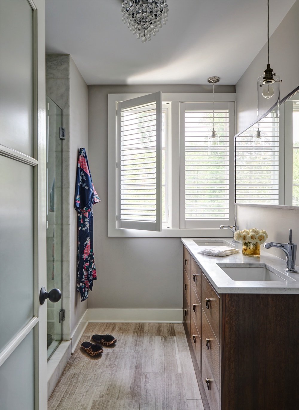 Full view of the storybook primary bathroom with walnut vanity, glass shower and slippers on the wood floor, by Martire Custom Homes