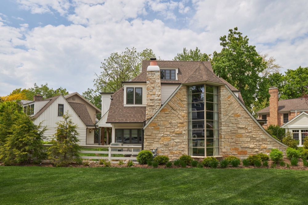 Rear exterior of a restored storybook home showing the full-height stone stair bay, curved rooflines and stone chimney, by Martire Custom Homes