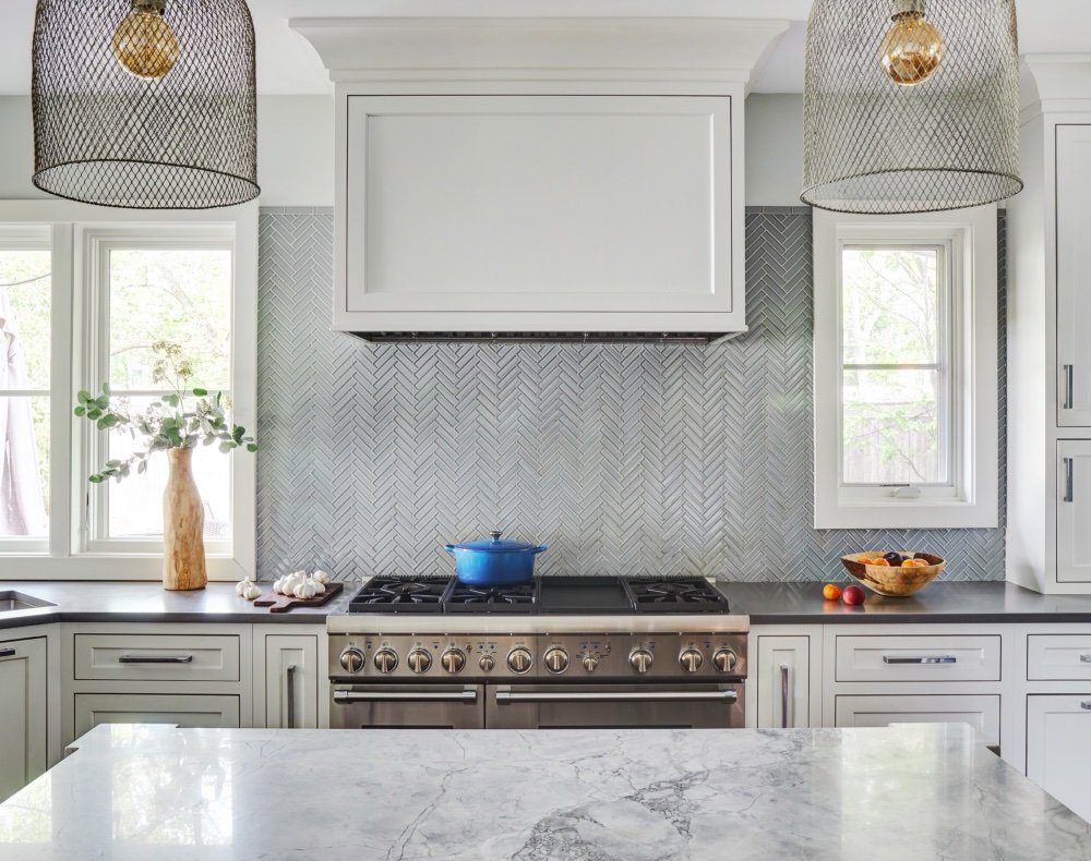 Storybook kitchen range wall with paneled hood, herringbone marble backsplash and woven pendants flanked by windows, by Martire Custom Homes