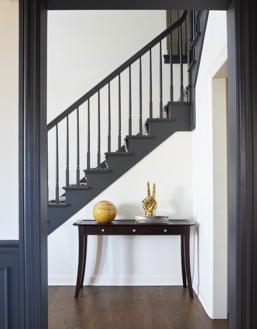 View through a charcoal-trimmed doorway to a dark painted staircase with turned balusters above a slim console table, by Martire Custom Homes