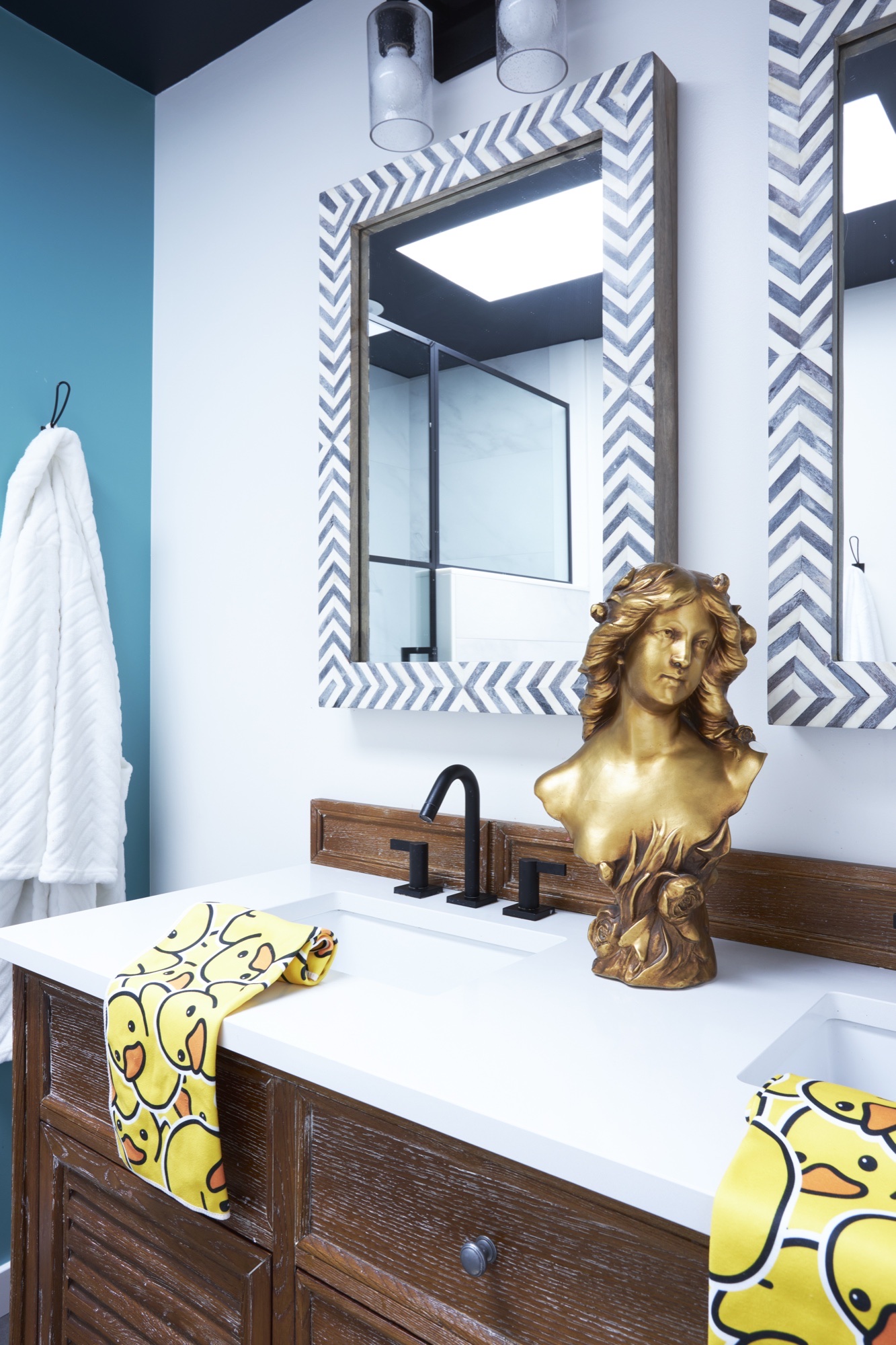 Angled view of a children's bathroom vanity with chevron-framed mirror, matte black faucet, teal accent wall and white robe on a hook, by Martire Custom Homes
