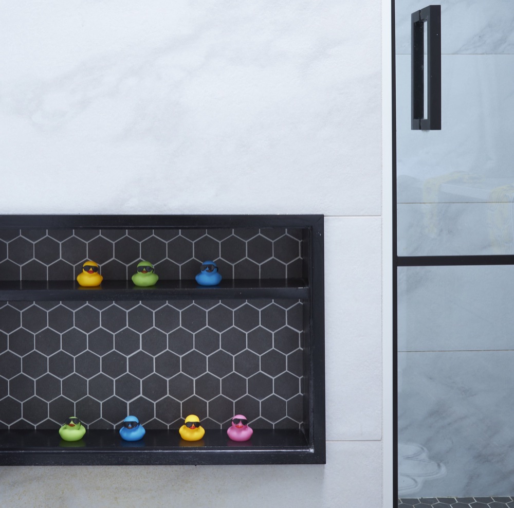 Black-framed shower niche lined with dark hexagon mosaic tile displaying colorful rubber ducks beside a black-framed glass shower door, by Martire Custom Homes