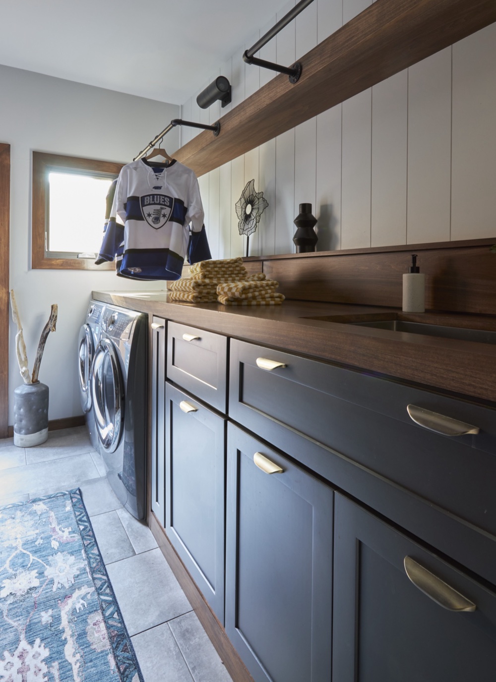 Laundry room run of charcoal cabinetry with brass pulls, walnut countertop and backsplash, shiplap walls and a blackened-pipe drying rail, by Martire Custom Homes