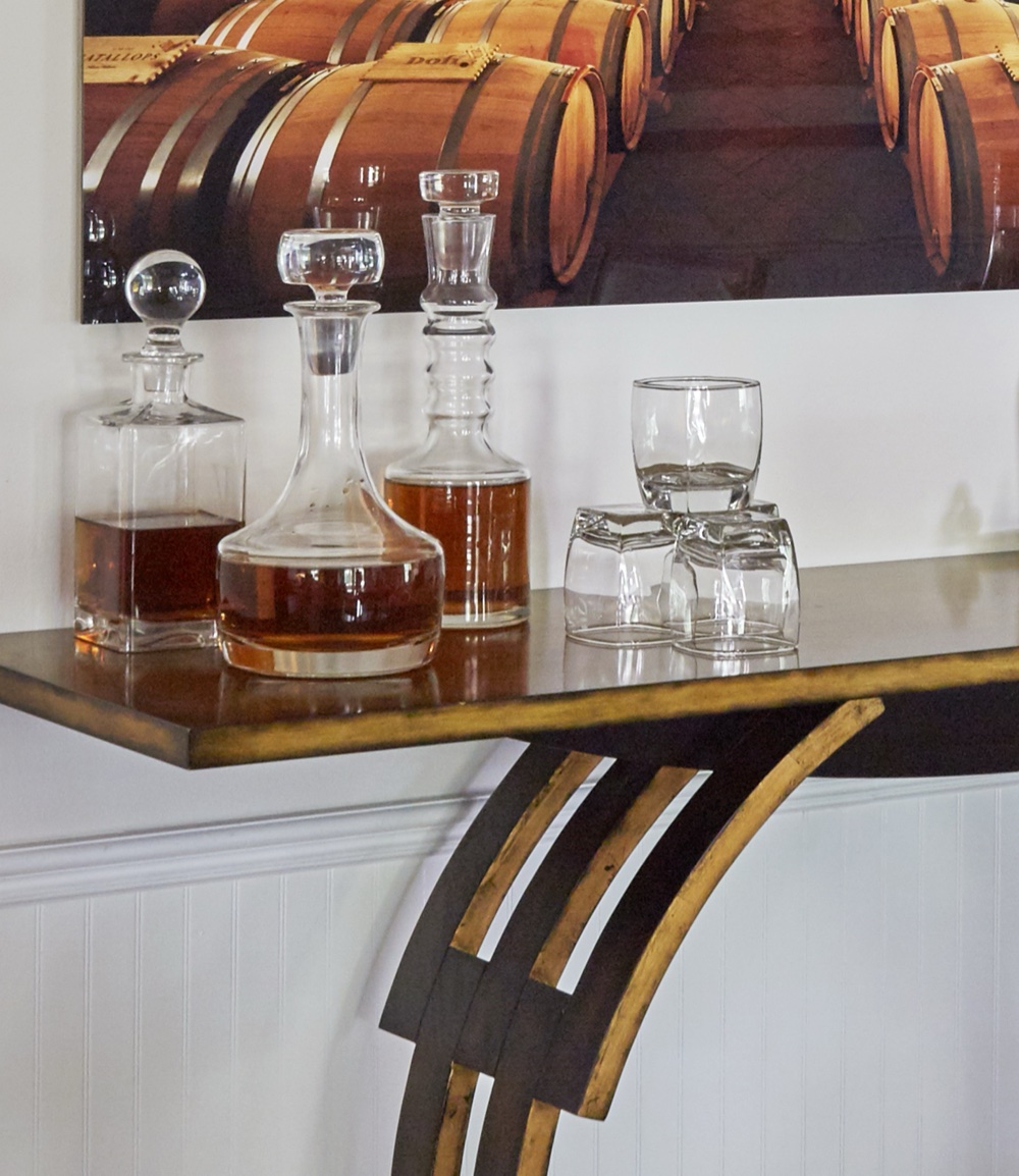 Bar vignette with crystal decanters and tumblers arranged on a gilt iron console against white beadboard wainscoting, by Martire Custom Homes