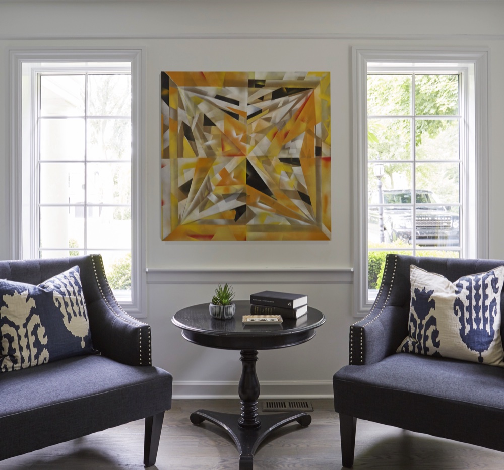 Sitting area with two navy nailhead-trimmed chairs flanking a black pedestal table beneath abstract artwork between tall windows, by Martire Custom Homes