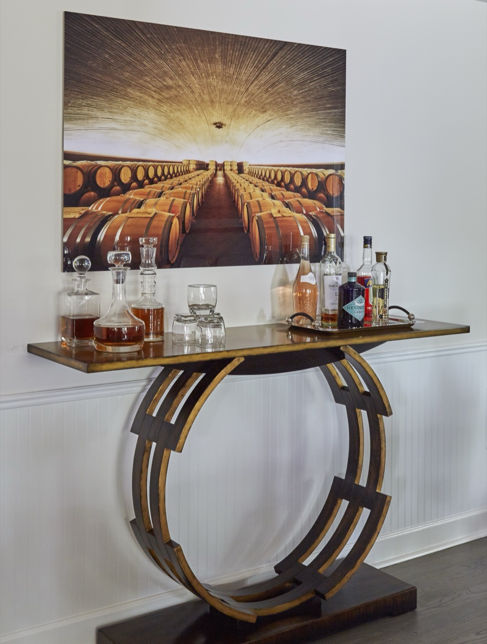 Bar console with a sculptural gilt ring base holding decanters and glassware beneath a large wine cellar photograph, by Martire Custom Homes