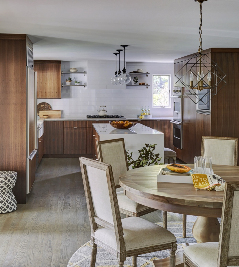 View from the dinette toward the kitchen showing rift-cut walnut cabinetry, a quartz waterfall island and a geometric brass chandelier over the round dining table, by Martire Custom Homes