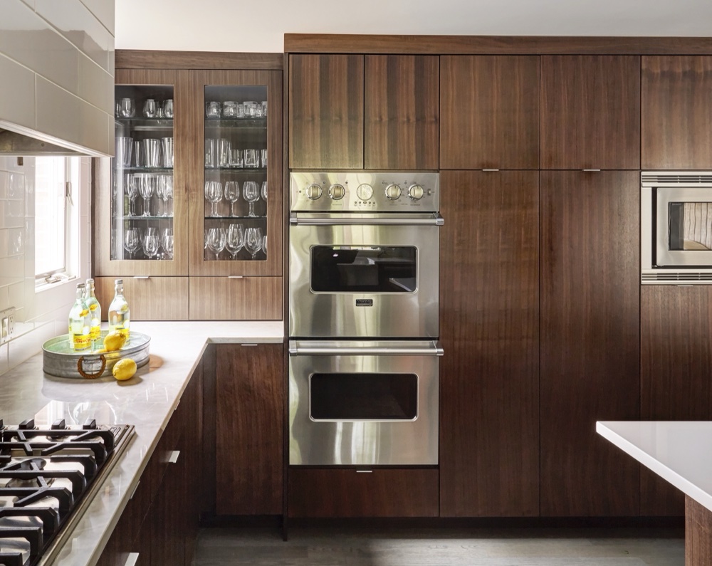 Floor-to-ceiling rift-cut walnut cabinetry with glass-front glassware display, stainless double wall ovens and built-in microwave, by Martire Custom Homes